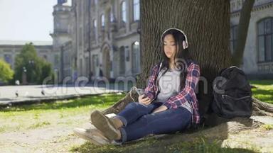 多种族女孩戴着耳机坐在树下，用电话，选择歌曲